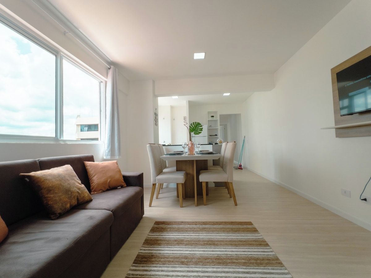 Una sala de estar moderna con un sofá, una mesa de comedor con sillas, un televisor montado en la pared y un ventanal que proporciona luz natural.