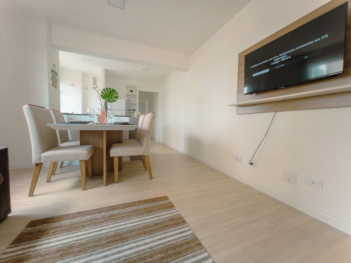 Ein modernes Wohnzimmer mit Flachbildfernseher an der Wand, einem Essbereich mit Holztisch und Stühlen sowie einer im Hintergrund erkennbaren Küche.