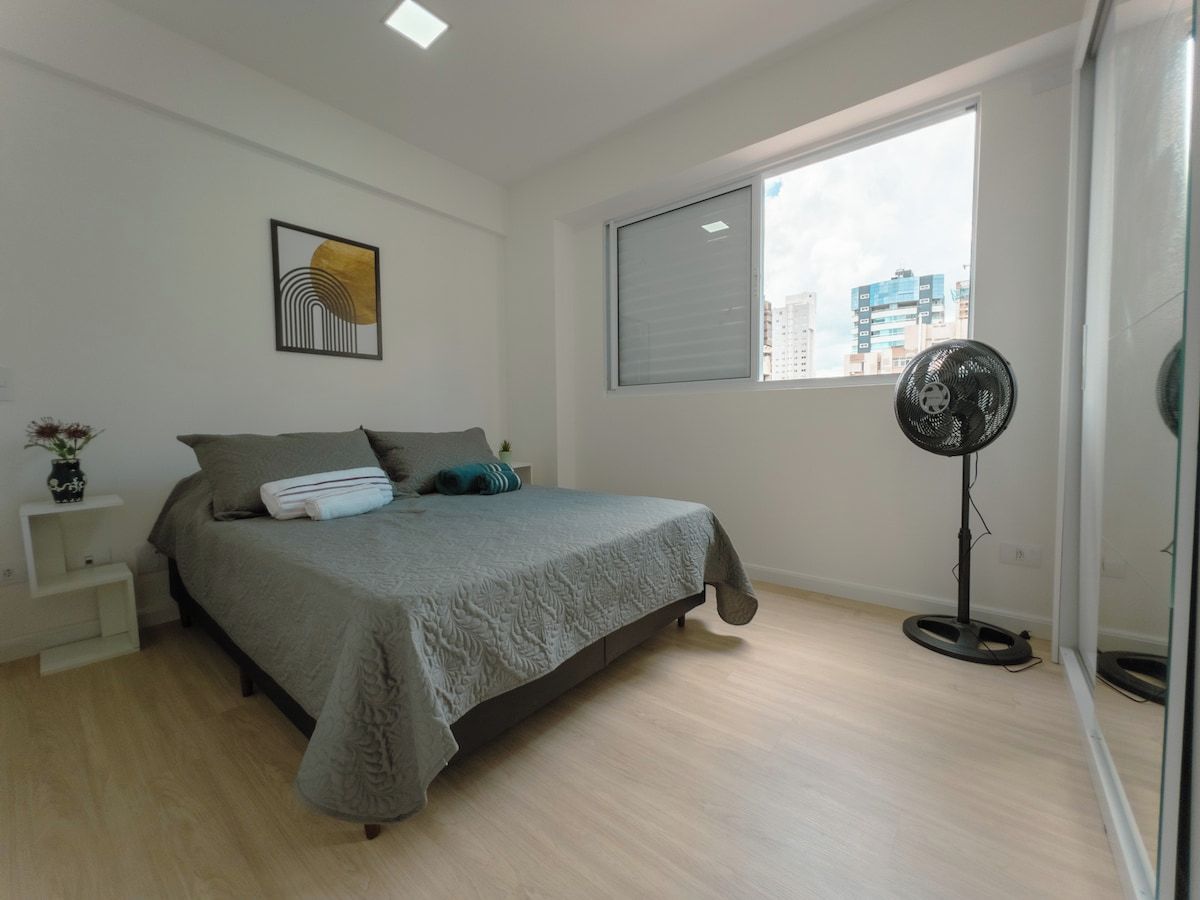 A bedroom with a bed, a bedside table, a floor fan, and a window with a view of buildings.