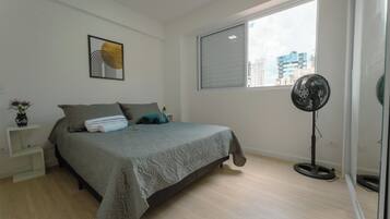 Un dormitorio con cama, mesita de noche, ventilador de pie y ventana con vistas a edificios.