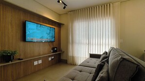 Un saló amb televisió de pantalla plana penjada a una paret de fusta, un sofà, una màquina de cafè i una finestra amb cortines.