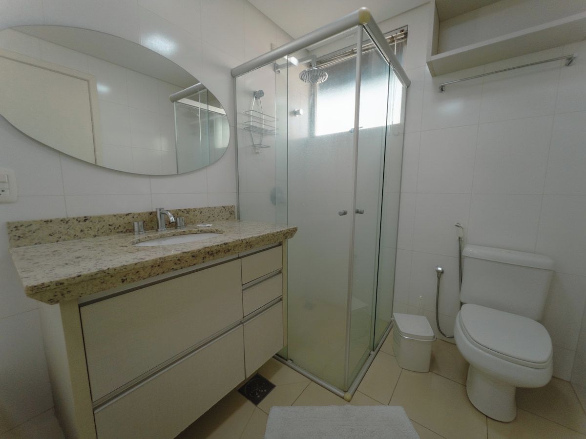 Casa de banho com chuveiro em cabine de vidro, sanita, lavatório com bancada de granito e espelho.