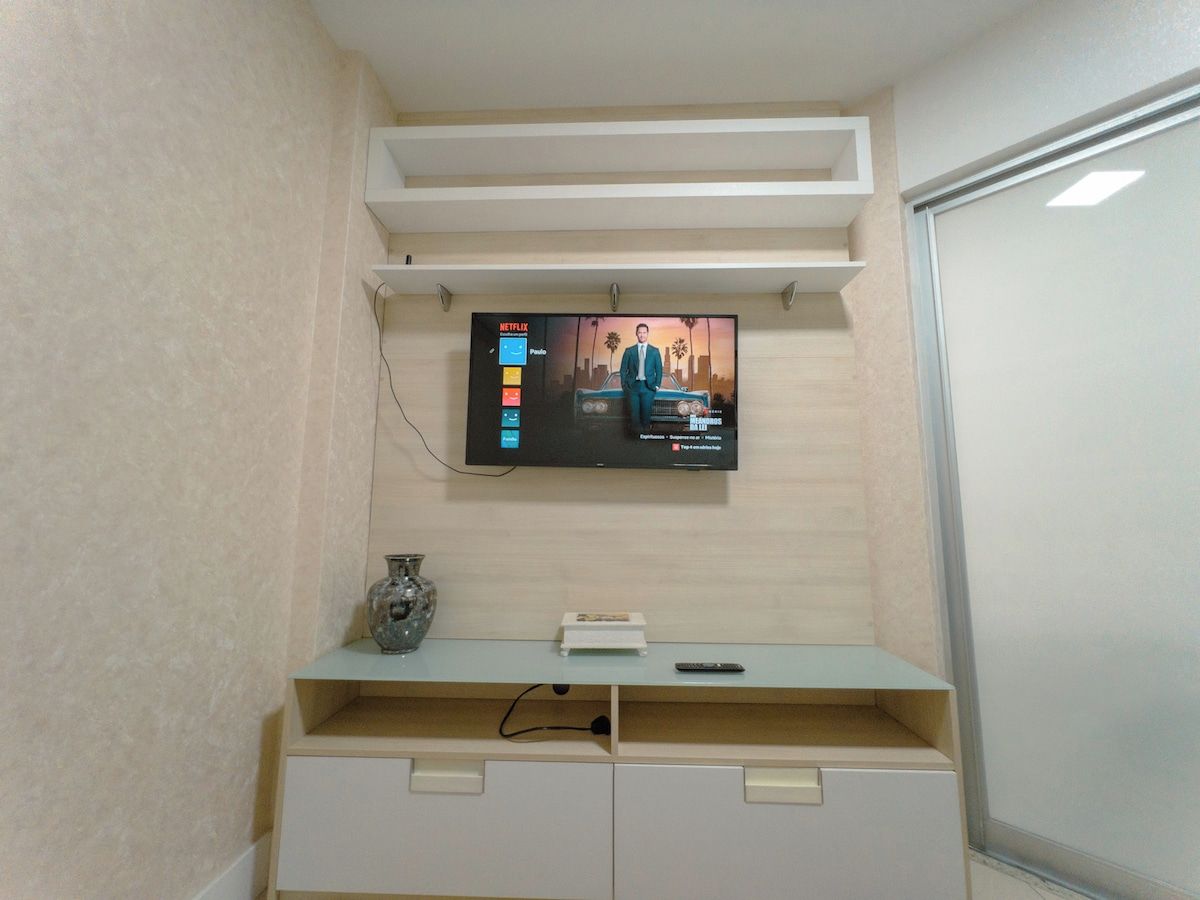 Un televisor montado en la pared con la interfaz de Netflix, un estante pequeño con un florero, y una puerta blanca.