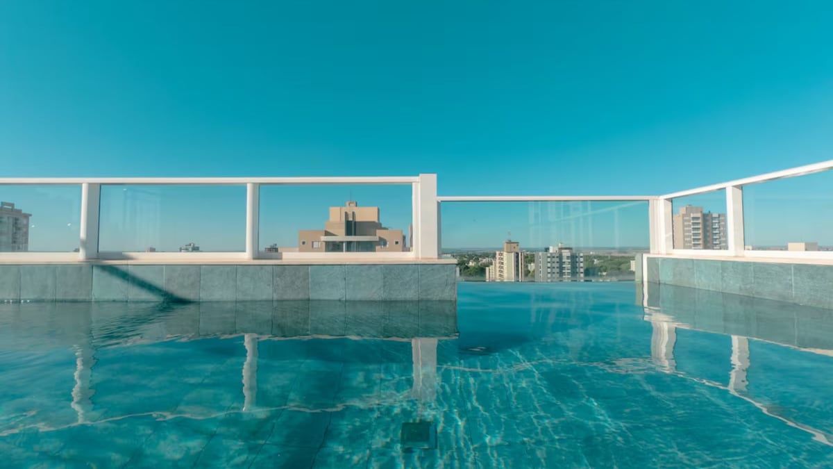 A rooftop swimming pool with clear blue water, surrounded by glass railings, and a view of distant buildings.