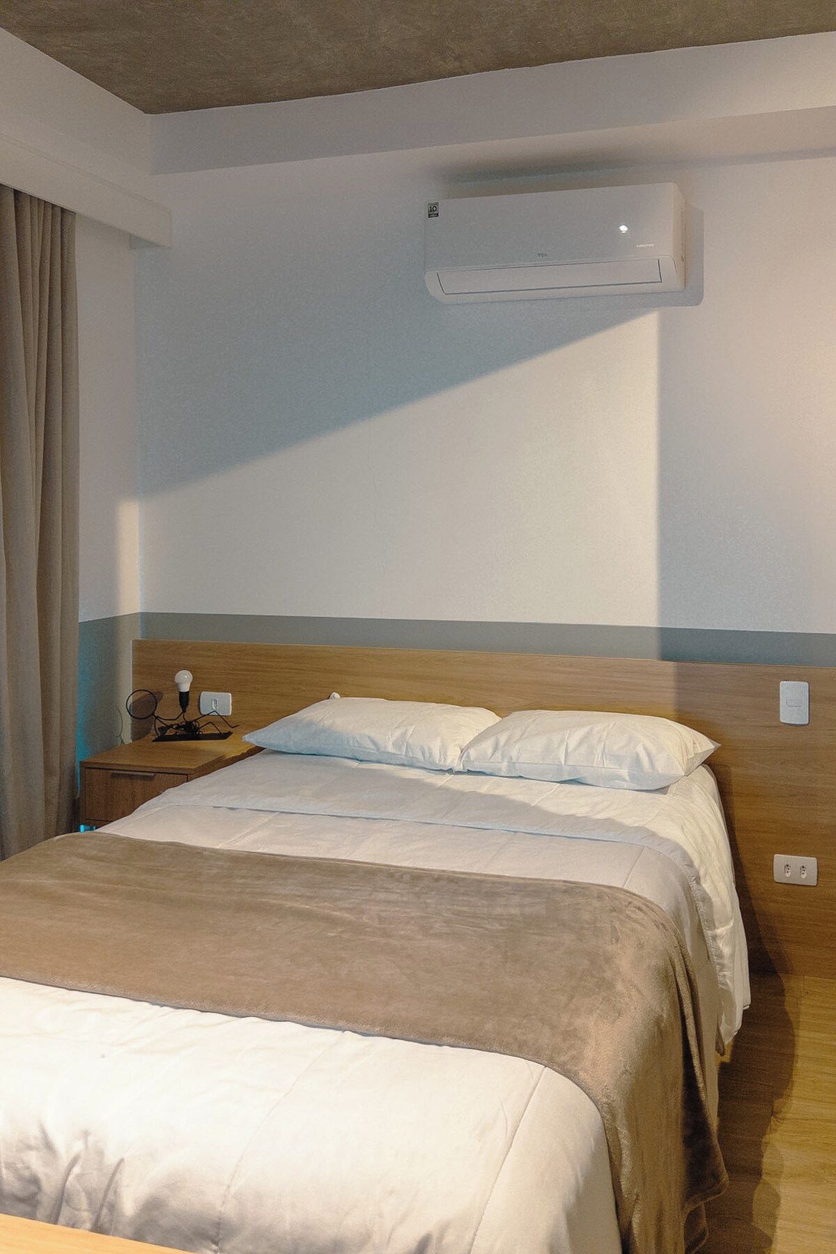 Quarto de hotel com cama, mesa de cabeceira, luminária e aparelho de ar condicionado.