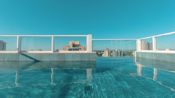 A rooftop infinity pool with clear blue water, surrounded by a white railing, and a view of distant buildings. - Private Room with Pool and Gym - BEDEO302B (Maringá)
