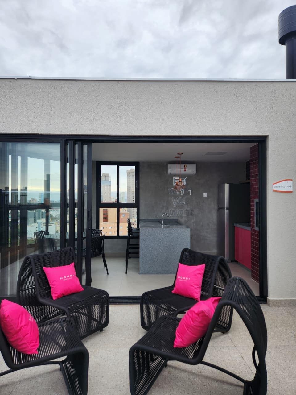 Un appartement moderne doté d’un balcon, d’une cuisine et d’un coin repas avec des meubles en rotin noir et des coussins roses.