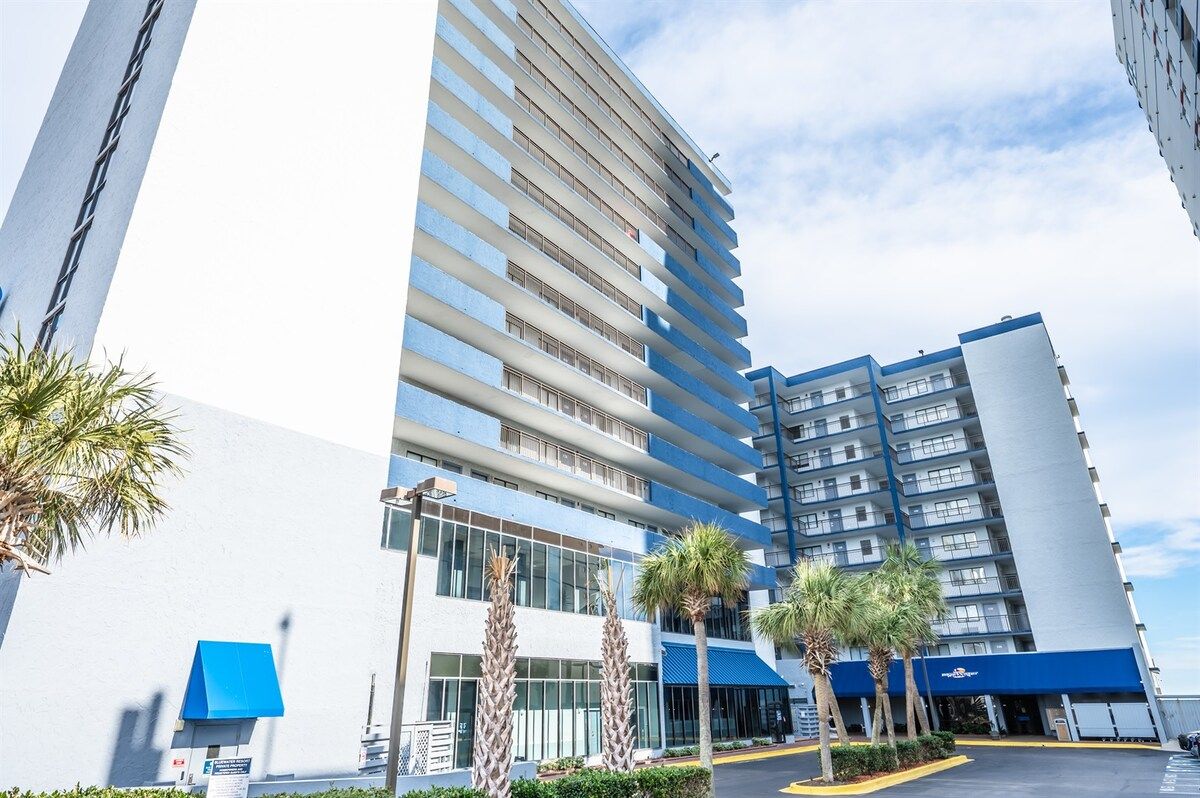 Un hotel de varios pisos con diseño moderno, que destaca por su esquema de color azul y blanco, y un estacionamiento con palmeras.