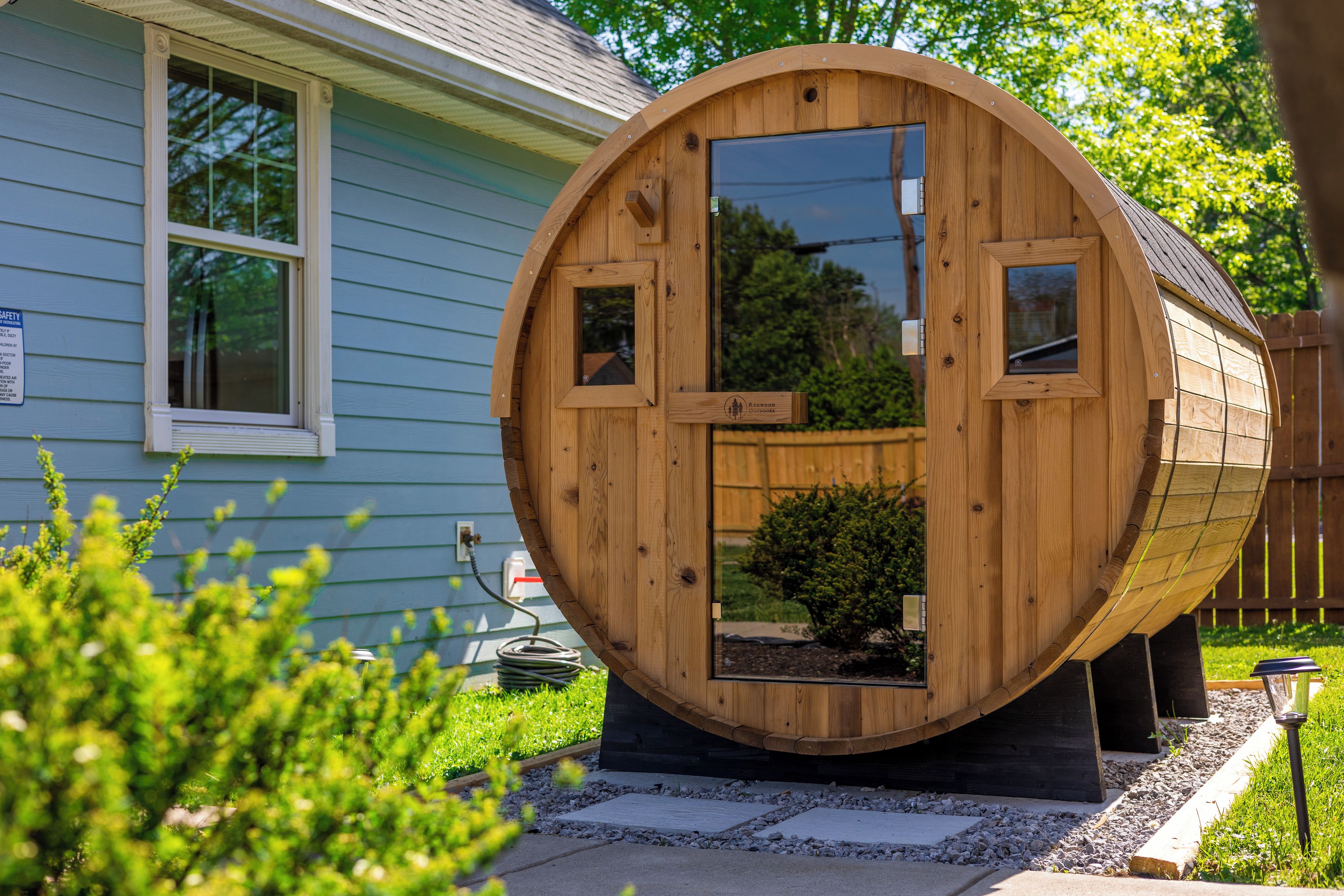 Una estructura de madera con forma de barril, con puerta y ventanas, sobre una base de hormigón y un pequeño jardín delantero.
