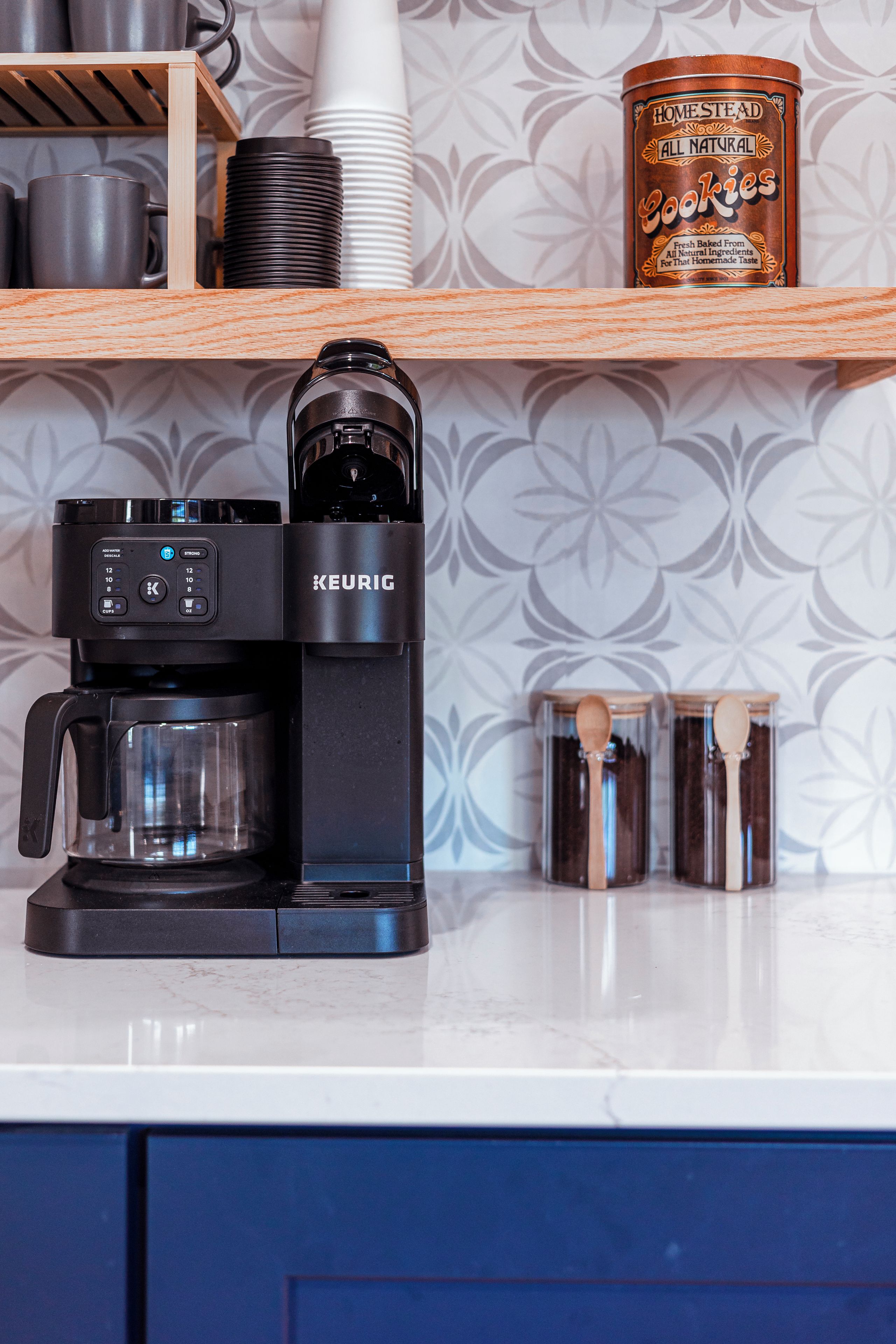 Una macchina da caffè Keurig su un bancone della cucina, con una caraffa, un barattolo di biscotti e un cucchiaio di legno.
