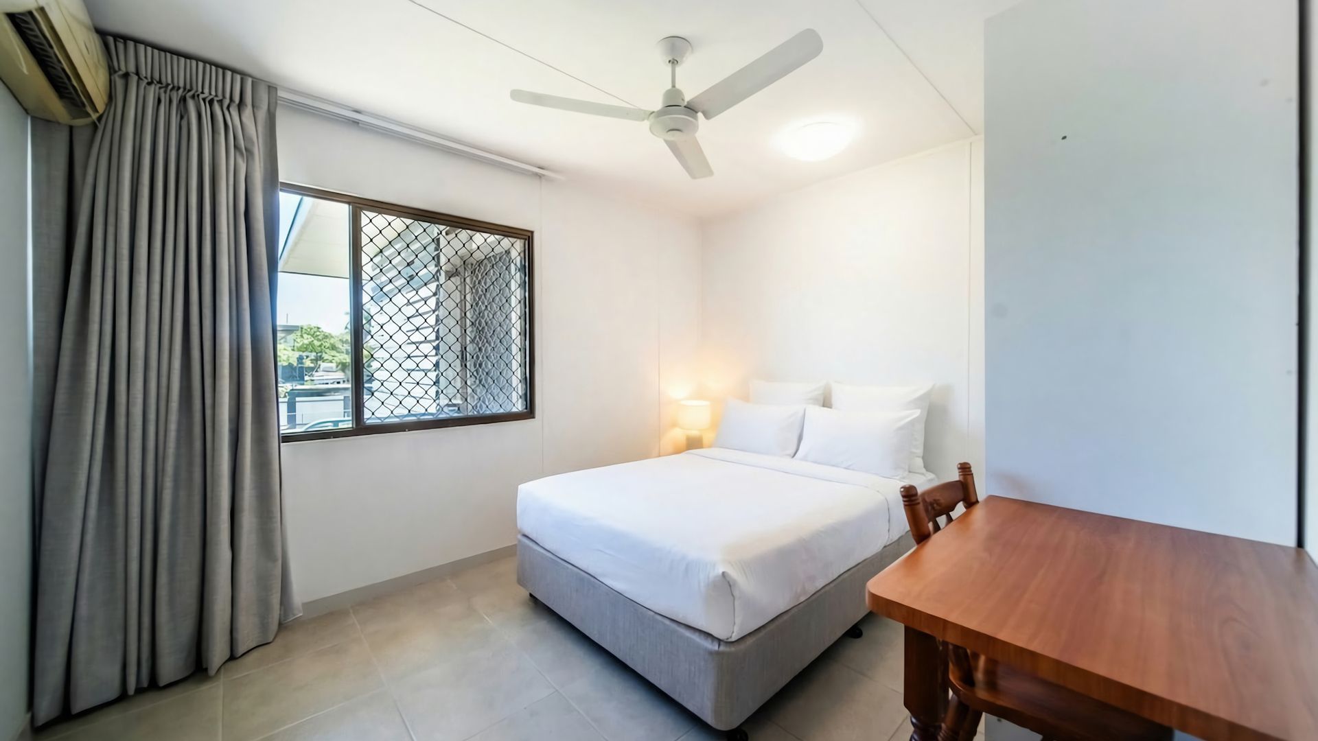 A hotel room with a bed, a table, a ceiling fan, and a window with curtains.