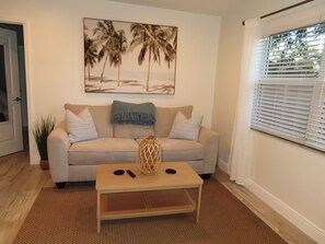 Een woonkamer met een beige bank, een salontafel, een schilderij van palmbomen, een plant en een raam met jaloezieën.