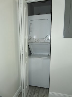 A compact laundry room with a front-loading washer and dryer, a door, and a light switch on the wall. - Linen and Palm 1bed/1bath heart of Fort Lauderdale (Fort Lauderdale)