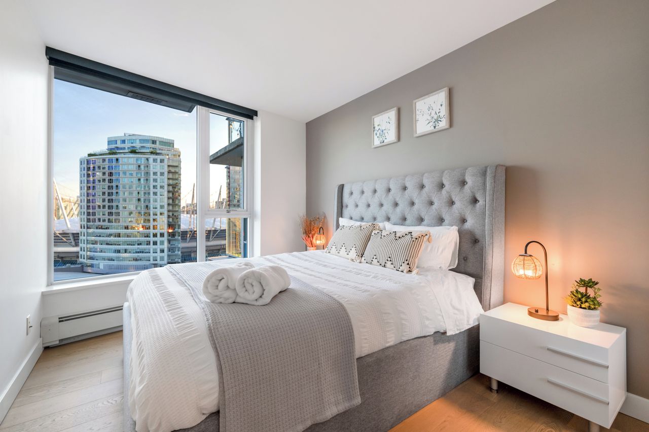 A bedroom with a large bed, a nightstand, a lamp, and a window with a city view.