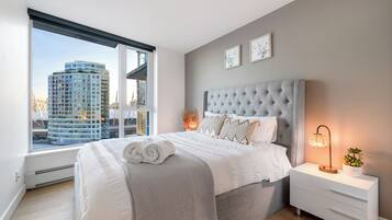 A bedroom with a large bed, a nightstand, a lamp, and a window with a city view.