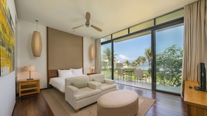 Ein modernes Hotelzimmer mit einem großen Bett, einer Couch, einem runden Hocker, einem Deckenventilator und einem Balkon mit Blick auf Palmen.