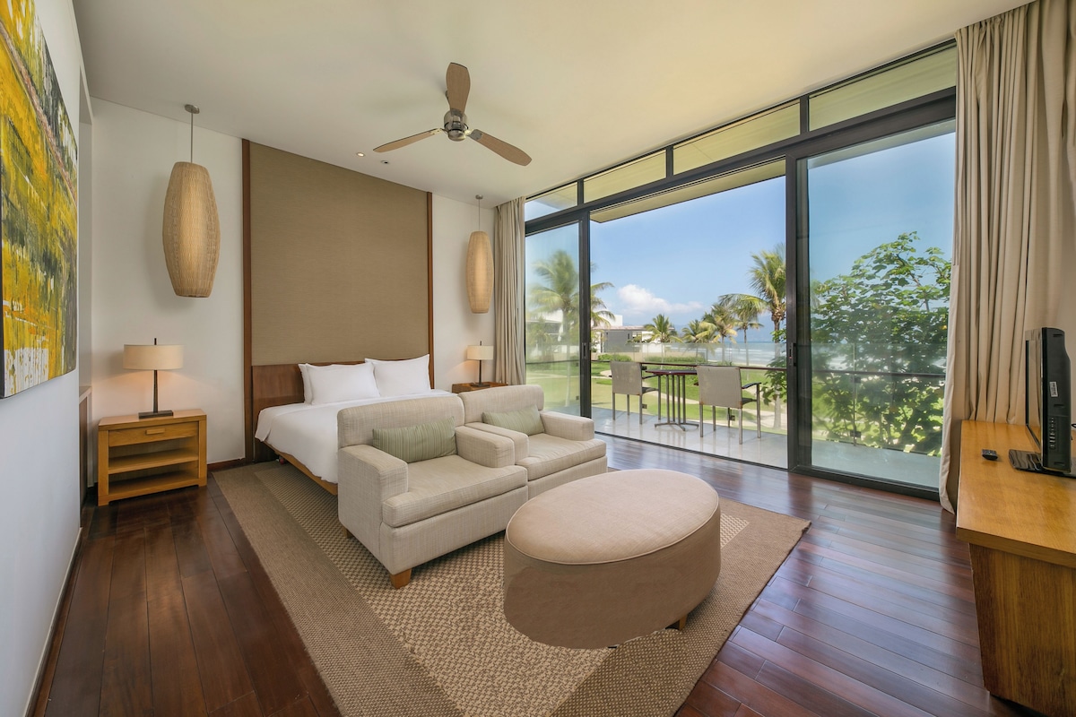 A modern hotel room with a large bed, a sofa, a round ottoman, a ceiling fan, and a balcony with a view of palm trees.