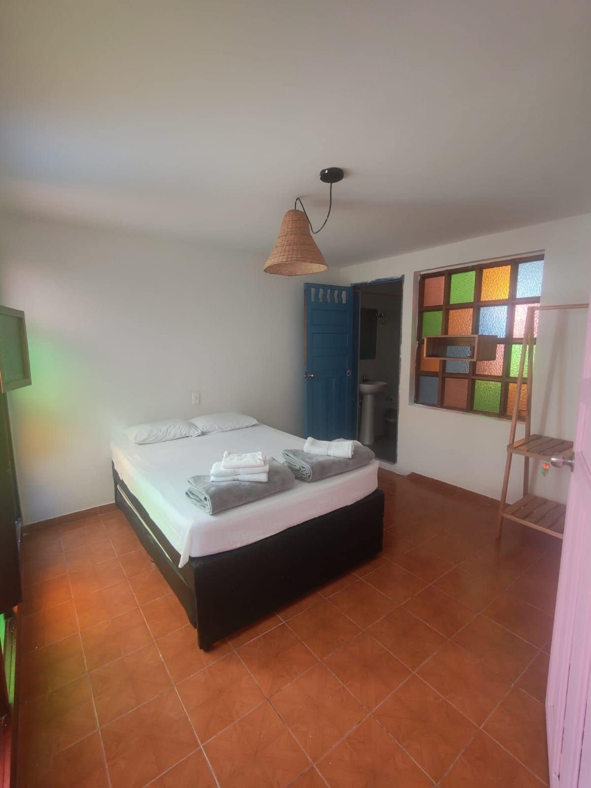 A bedroom with a bed, a blue door, a window with colored glass, and a hanging lamp.