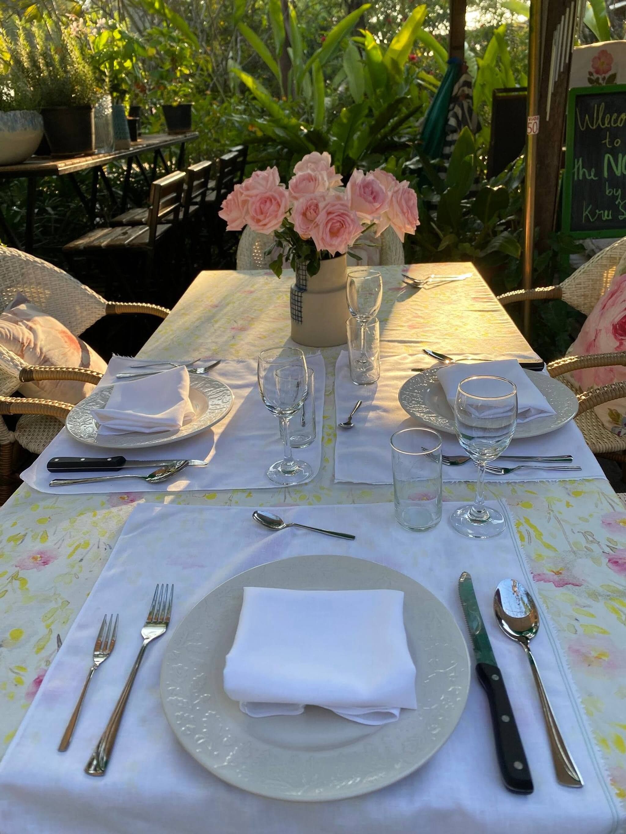 Ett bord dukat för middag utomhus, med en blommig duk, vita tallrikar, bestick och en vas med rosa blommor.