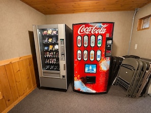 Two vending machines, one Coca-Cola branded and one with various snacks, in a room with wooden paneling and stacked chairs.