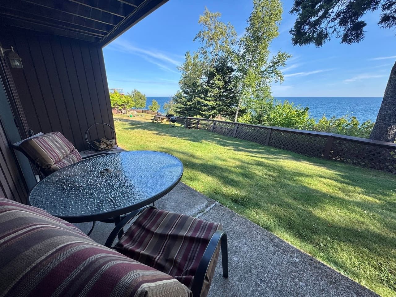 Une terrasse extérieure avec une table ronde et des coussins à rayures, donnant sur une pelouse et un plan d’eau.