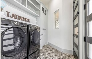 A laundry room with two front-loading washers and dryers, a white wall with a 'Laundry' sign, patterned floor tiles, and a door with a window. - Modern/Luxury/Rooftop Deck/Downtown/Heated Pool/5BDRM (Houston)