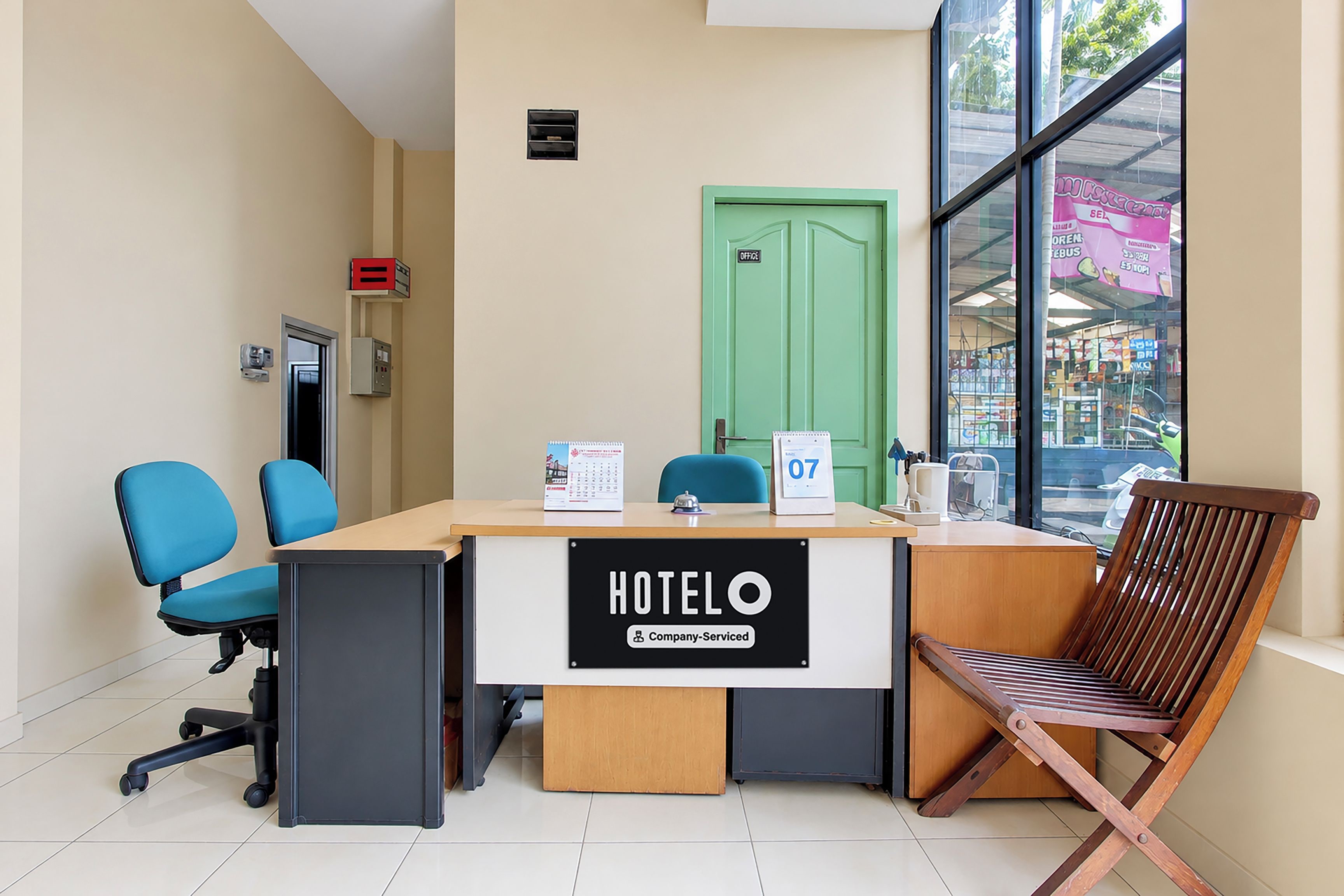 A hotel reception desk with a sign indicating it's company-serviced, a green door labeled 'office', and a view of a street through large windows.