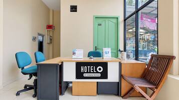 A hotel reception desk with a sign indicating it's company-serviced, a green door labeled 'office', and a view of a street through large windows.