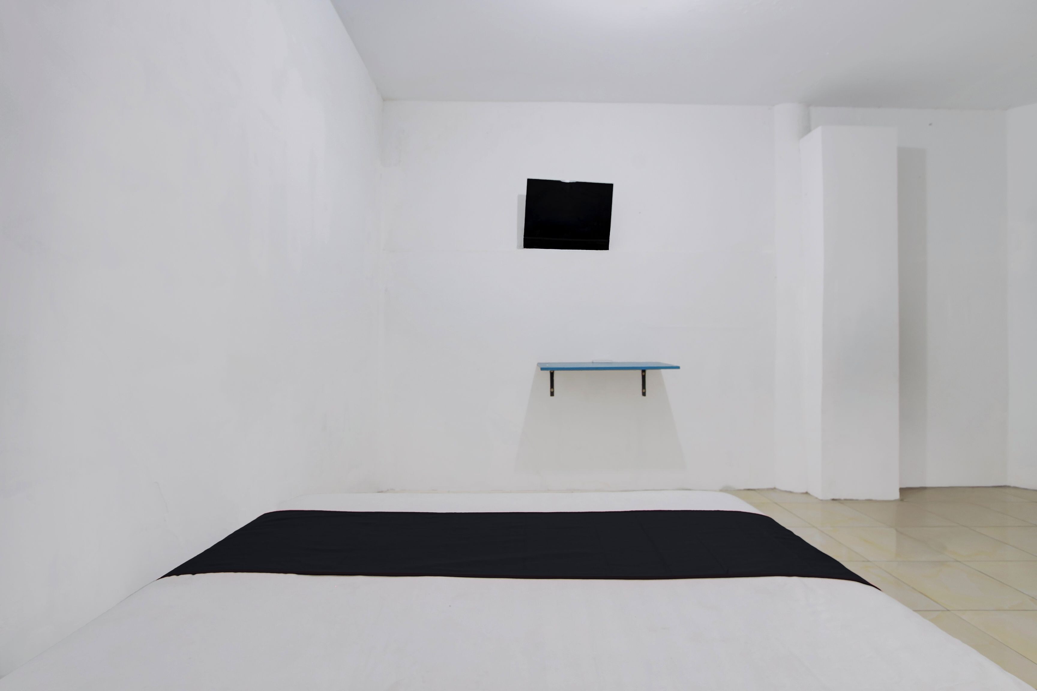 A minimalist room with a bed, a small blue shelf, and a black object on the wall.