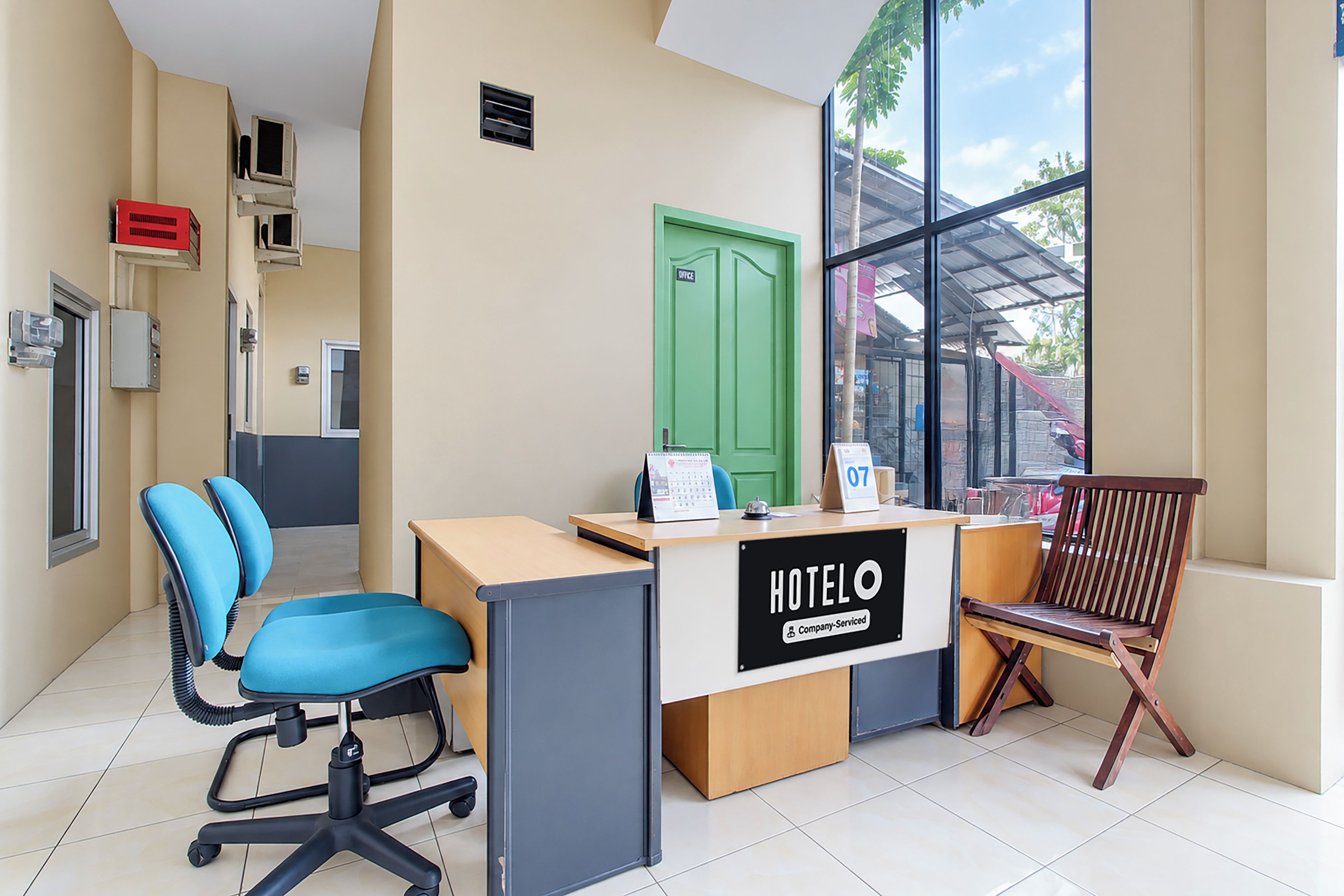 Ein Hotelrezeptionstresen mit einem Schild, das die Firmenbetreuung kennzeichnet, eine grüne Tür mit der Aufschrift 'Office' und ein Blick ins Freie durch große Fenster.