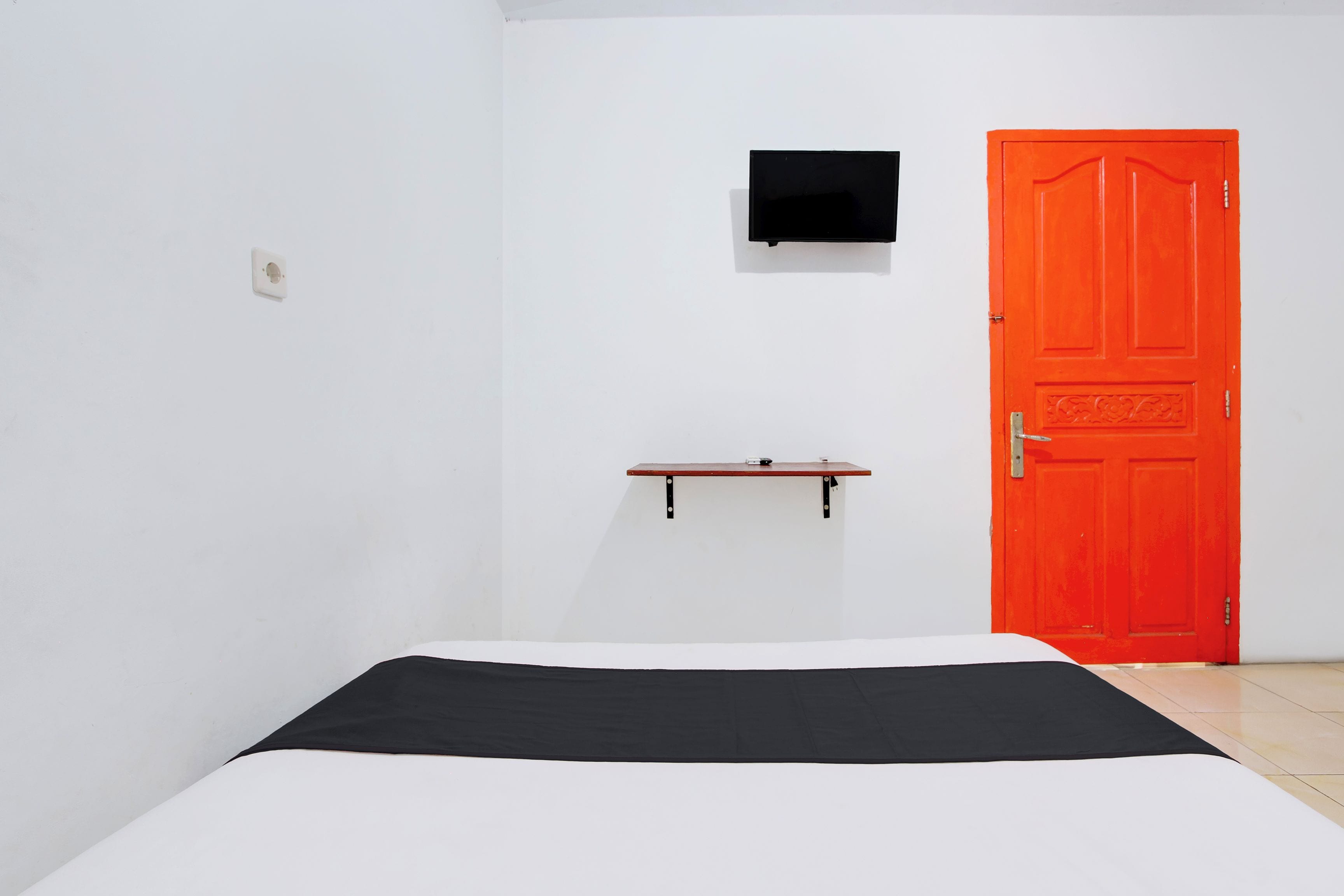 A room with a bed, a red door, a small shelf, and a wall-mounted TV.