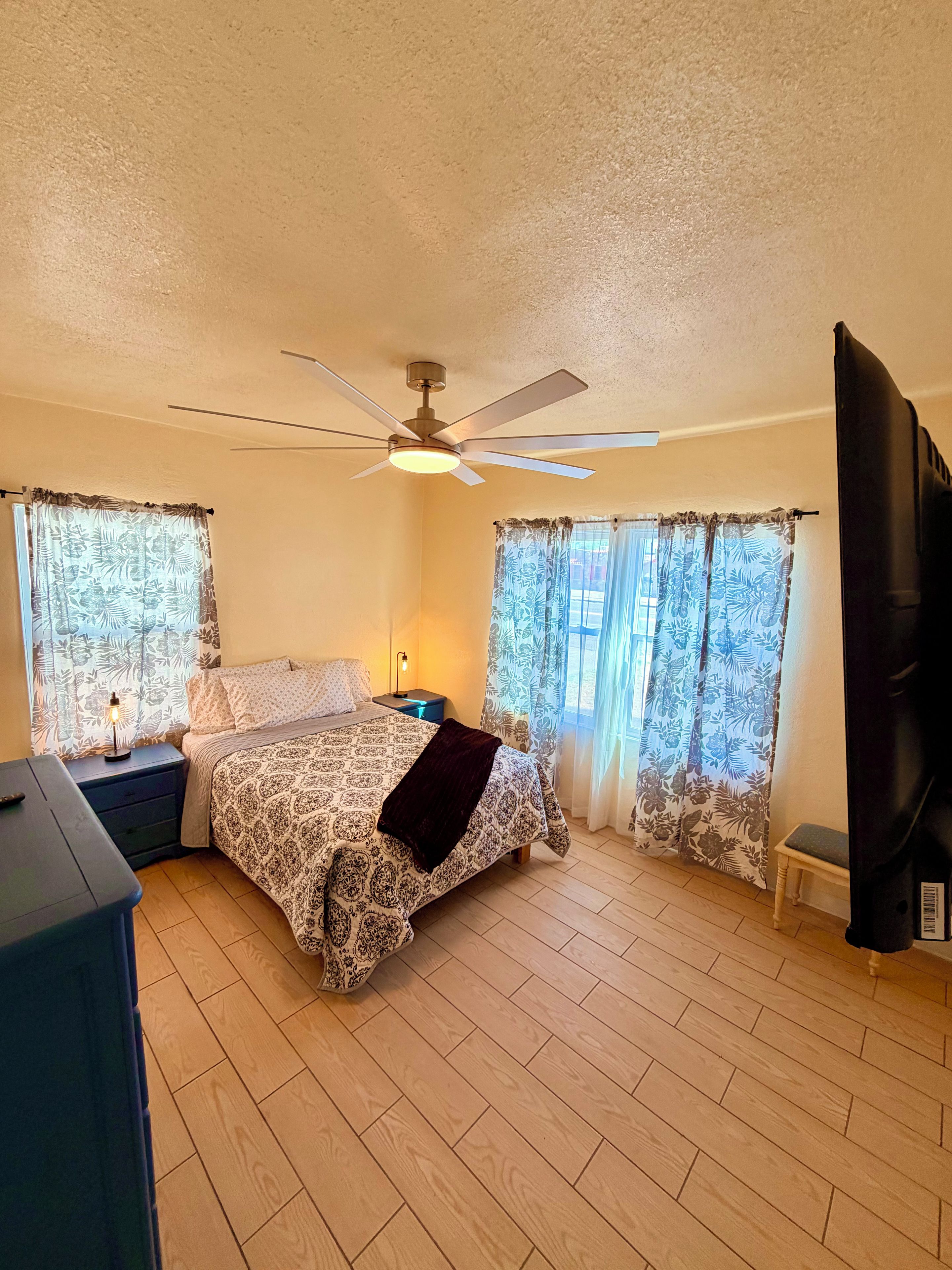 A bedroom with a bed, a ceiling fan, a nightstand with a lamp, and a window with curtains.