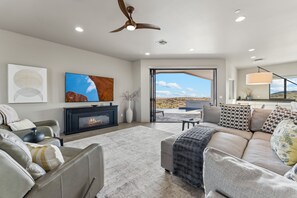 A modern living room with a fireplace, comfortable seating, and a large window offering a view of the desert landscape. - The Mountain Watch | Infinity Pool & Casita (Fountain Hills)