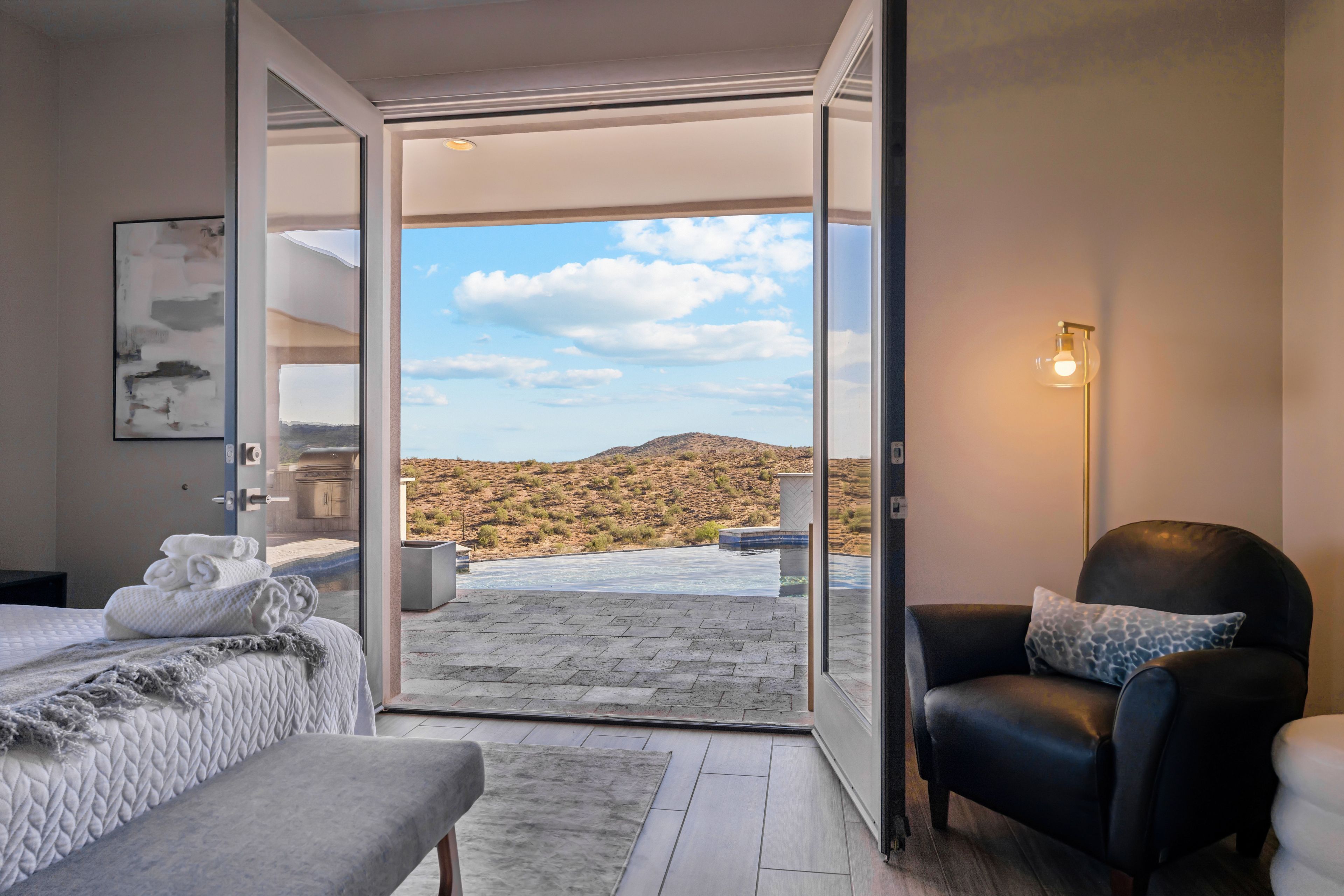 Habitación de hotel con cama, una silla, un vidrio que da a una piscina y vistas de un paisaje desértico.
