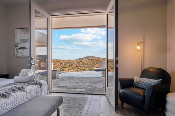 Quarto de hotel com cama, uma cadeira, porta de vidro para piscina e vista para paisagem desértica.