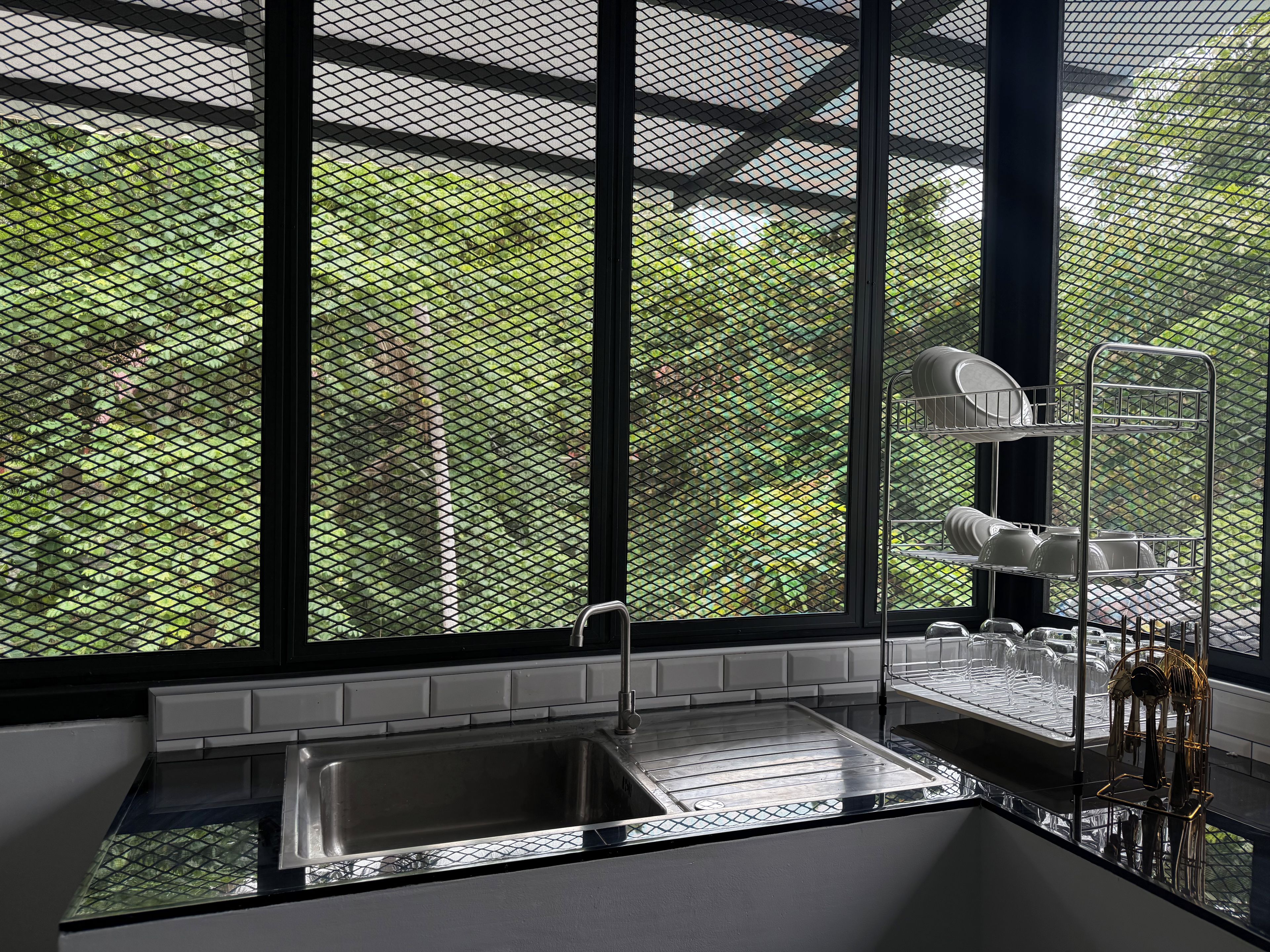 A modern kitchen with a stainless steel sink, a dish rack, and a view through a mesh window to a green outdoor area.