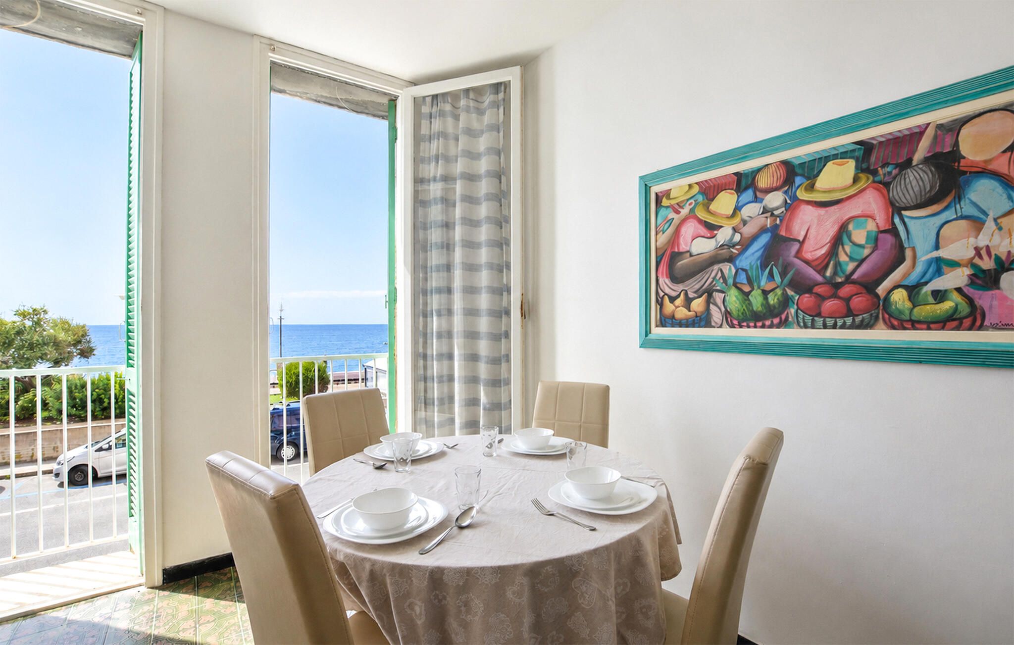 Un coin repas avec une table dressée pour quatre personnes, une vue sur la mer par une porte ouverte et un tableau coloré accroché au mur.