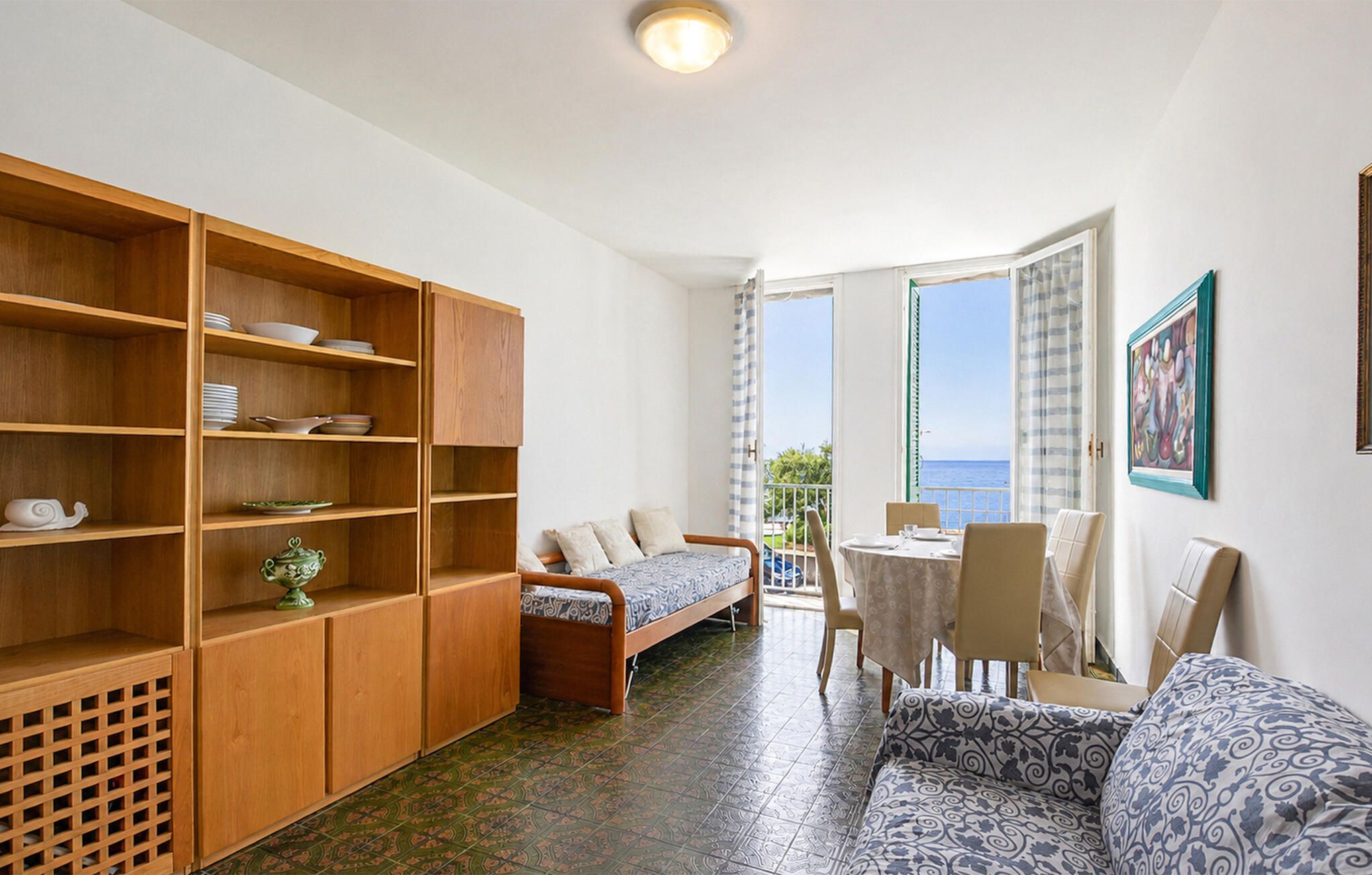 A room with a bed, a sofa, a dining table with chairs, a wooden cabinet with dishes, and a view of the sea through a window.