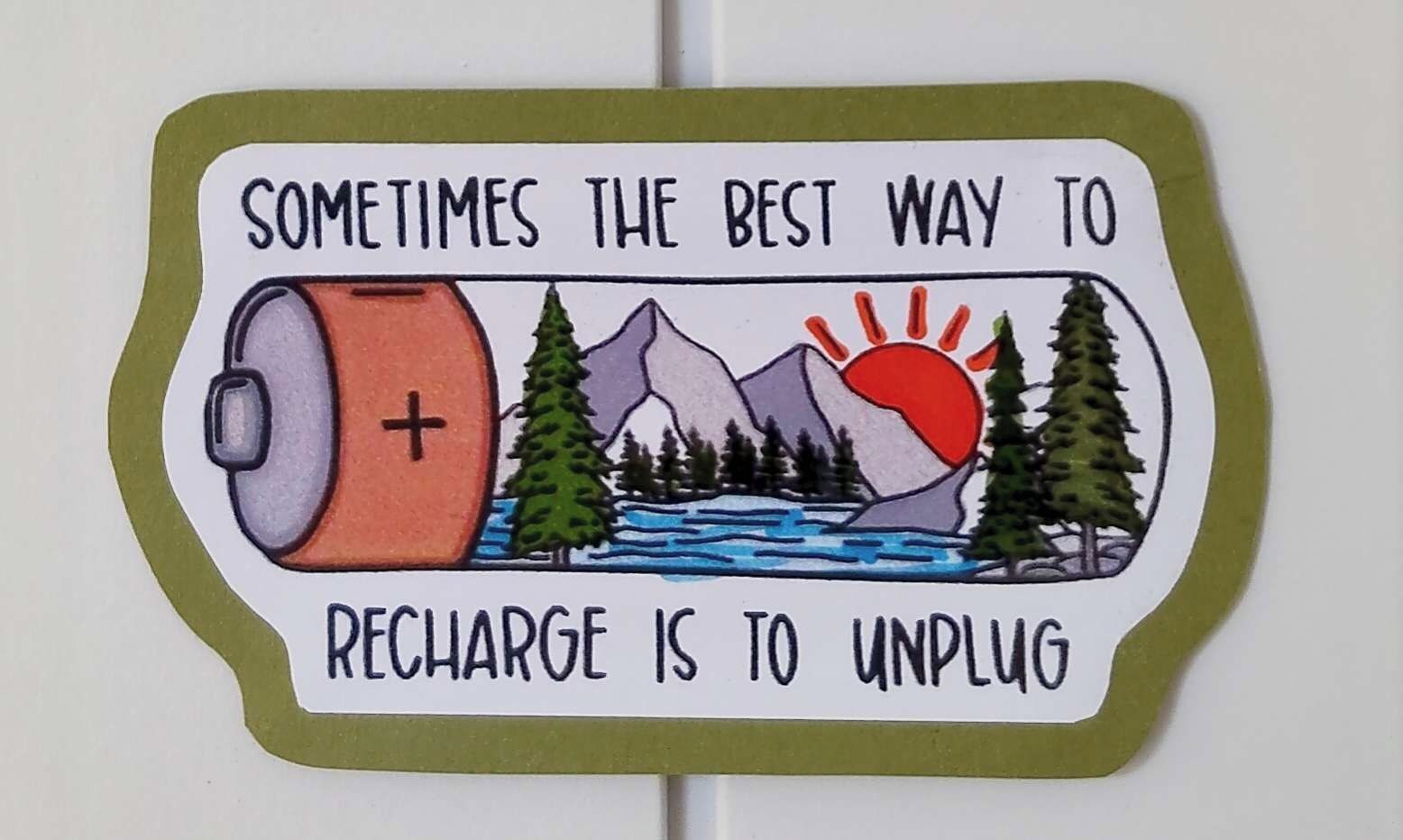 Ein Aufkleber mit einer Berglandschaft und einer Akku-Illustration, mit dem Text 'Manchmal ist der beste Weg, um neue Energie zu tanken, abzuschalten'.
