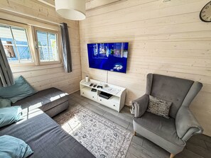 Ein gemütliches Wohnzimmer mit einem an der Wand befestigten Flachbildfernseher, einem grauen Sofa, einem gemusterten Teppich und einer weißen Unterhaltungsanlage mit Spielcontrollern.