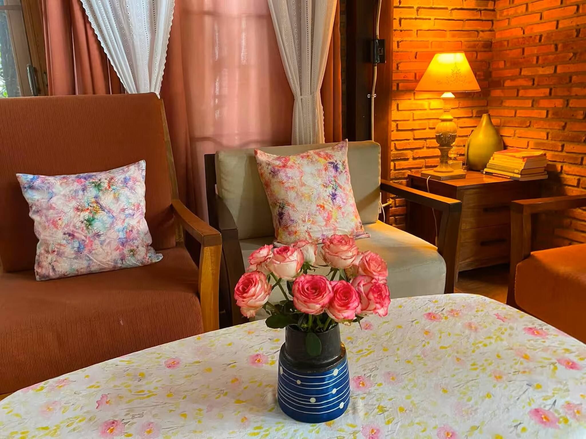 Et hyggeligt rum med en murstensvæg, et bord med dug med blomster, en vase med pink roser og to stole med blomsterpuder.
