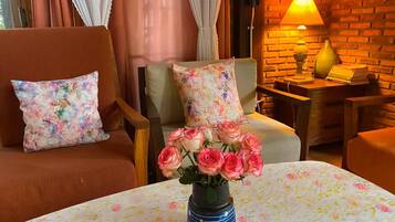 Et hyggeligt rum med en murstensvæg, et bord med dug med blomster, en vase med pink roser og to stole med blomsterpuder.