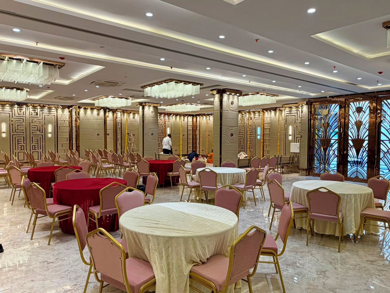 A banquet hall with round tables and chairs, a person in the background, and decorative wall panels.