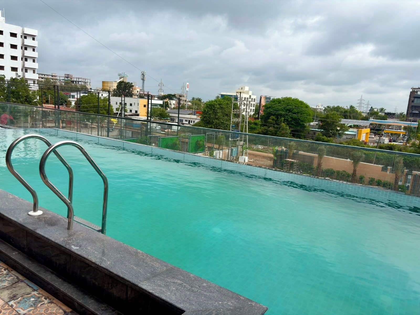 A swimming pool with a metal railing, surrounded by a fence, with buildings and trees in the background.
