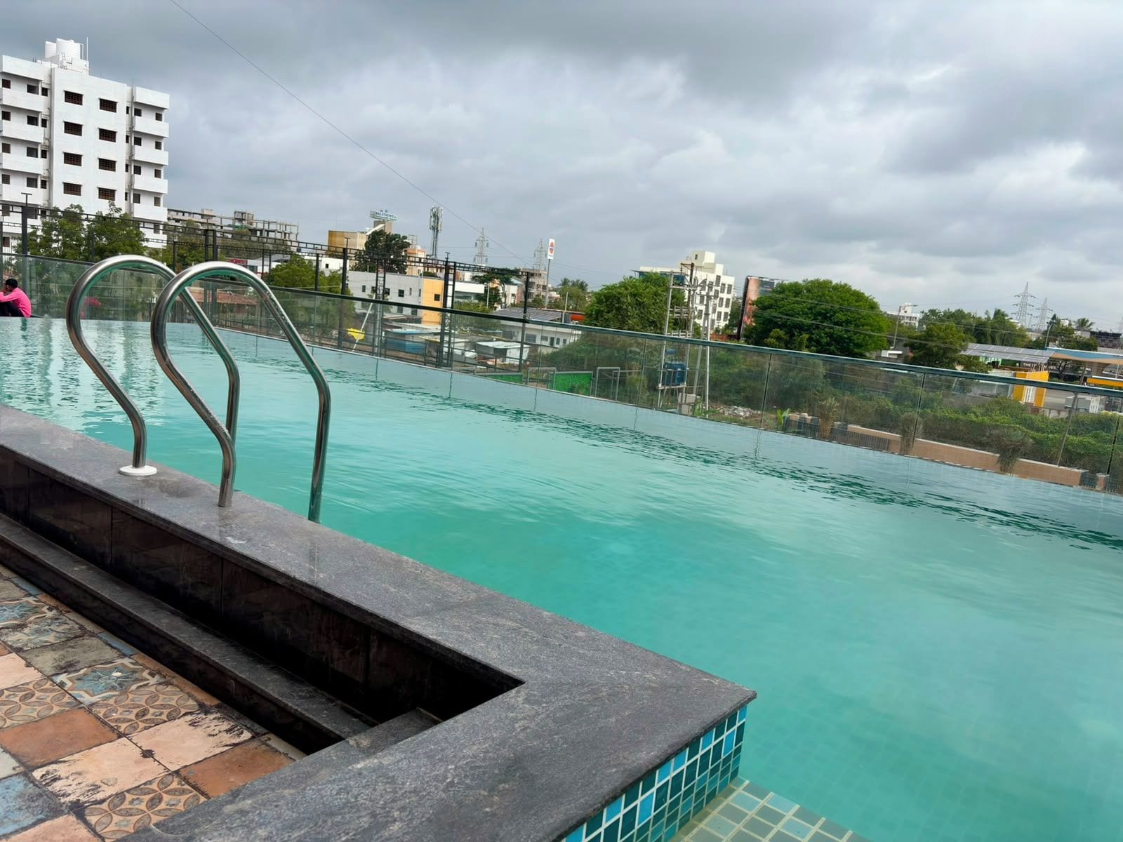 A swimming pool with a metal railing, a tiled deck, and a view of buildings in the background.
