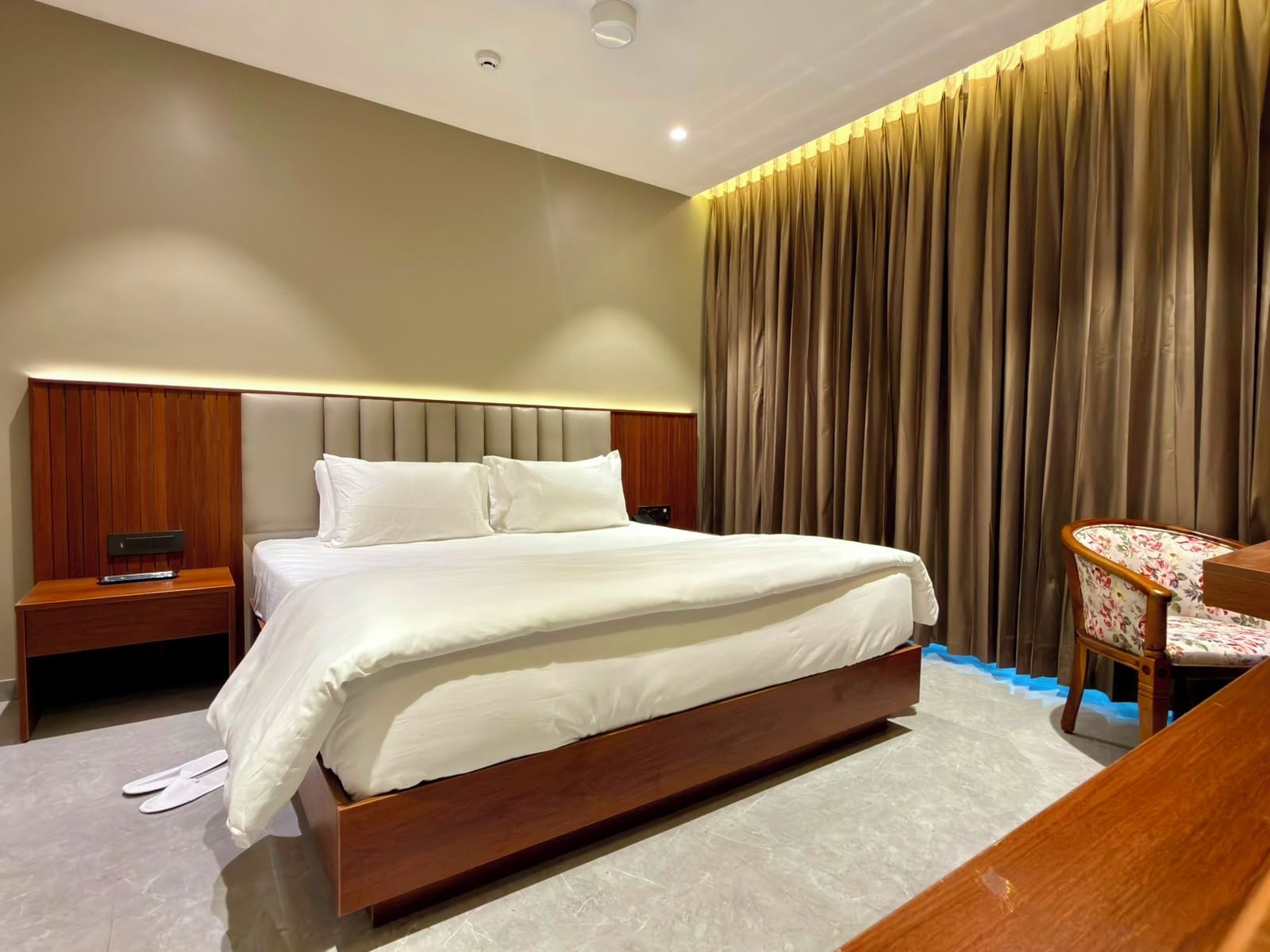 A modern hotel room with a large bed, a wooden nightstand, a desk with a chair, and a curtain backdrop.
