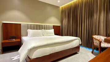 A modern hotel room with a large bed, a wooden nightstand, a desk with a chair, and a curtain backdrop.
