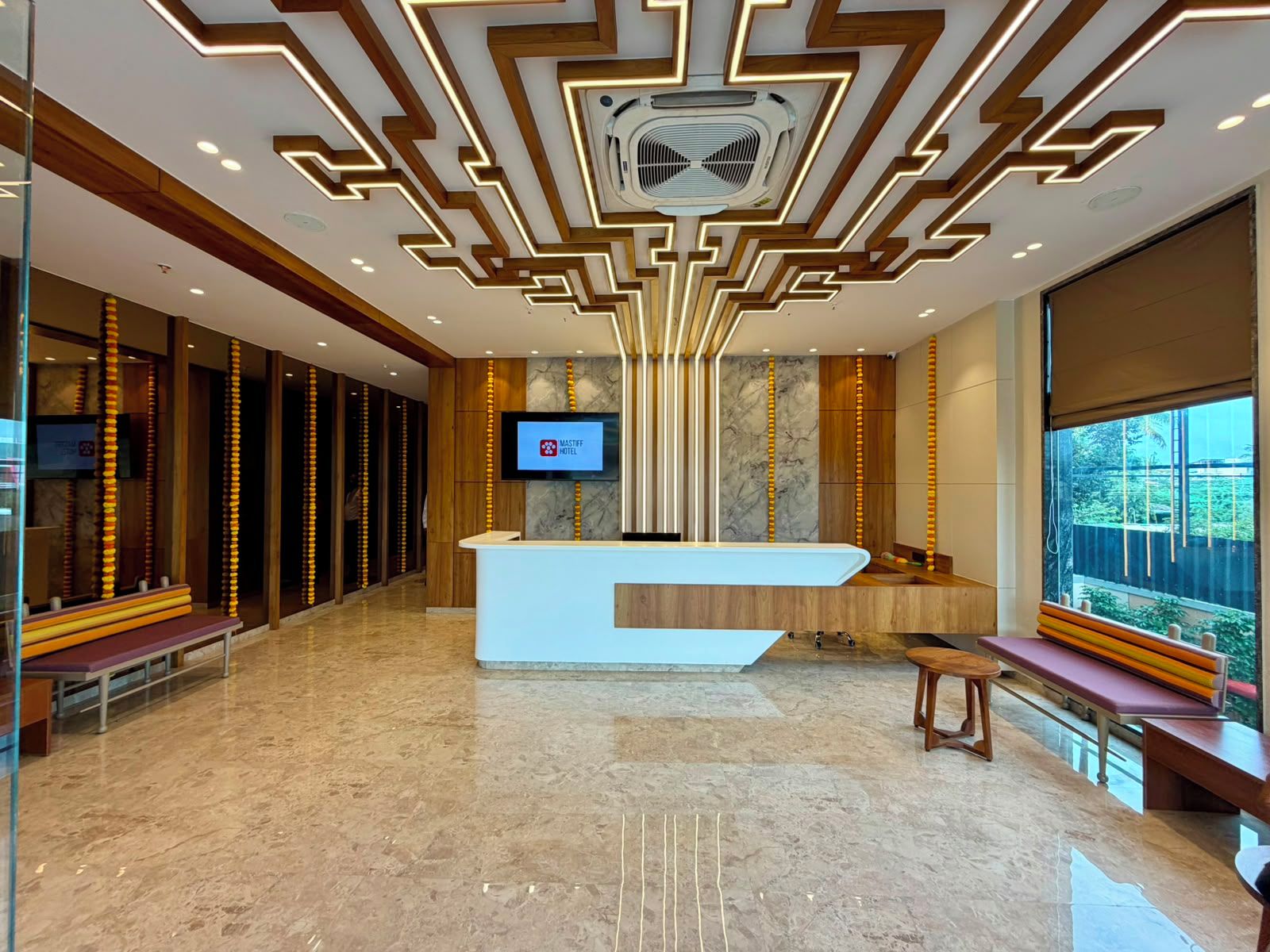 A modern hotel lobby with a reception desk, a television screen, and a large window offering a view of the outdoors.