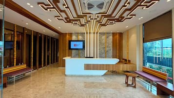 A modern hotel lobby with a reception desk, a television screen, and a large window offering a view of the outdoors.
