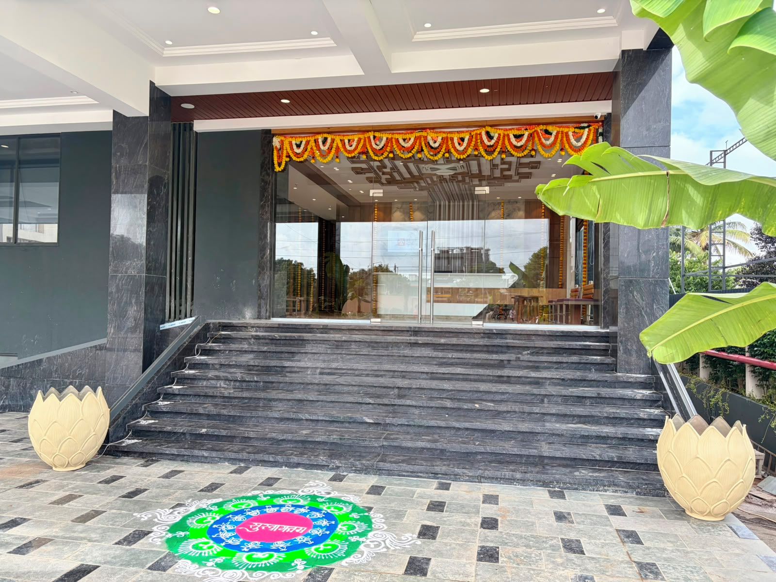 A modern building entrance with a staircase leading to a glass door, a decorative garland above the door, and a traditional rangoli design on the ground.